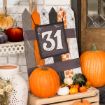 A festive Halloween display with pumpkins, gourds, and a sign reading "31 October" decorated with spiders, webs, bats, and pumpkins, set against a white brick wall and wooden door.
