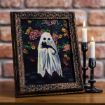A framed painting of a ghost holding a black cat, set against a floral background. The frame is ornate and sits on a wooden surface next to two lit, partially melted candles in black holders. A brick wall is in the background.