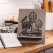 A rustic wooden recipe stand with a fall-themed design featuring pumpkins and a black bird, holding an open cookbook on a kitchen counter with eggs, a mixing bowl, and rolling pins in the background.