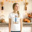A girl stands indoors wearing a white T-shirt with a scarecrow print. Behind her are pumpkins, autumn leaves, and corncobs on a wooden shelf against a white brick wall decorated with string lights shaped like leaves.