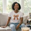A smiling woman sits on a couch wearing a white T-shirt with a large brown bow graphic. She has curly hair and light blue jeans. A bowl of popcorn is on the table in front of her, and soft natural light fills the room.