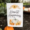 A garden flag with "Harvest Happiness" written in script, featuring sunflowers and pumpkins in orange, white, and green. The flag is displayed outdoors near pumpkins and soil.