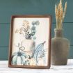 A framed artwork of a rustic vase with flowers, eucalyptus, and assorted blue and white pumpkins sits on a white table. Beside it is a gray clay vase holding dried wheat stalks, set against a teal wooden background.
