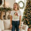 A smiling woman stands in a festive living room decorated for Christmas, wearing a white t-shirt with a jolly Santa face and the words "Feelin Jolly." A decorated tree and stockings are visible in the background.