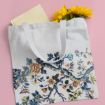 A white tote bag with floral and branch designs, holding a closed book and a bunch of yellow sunflowers, is placed on a pink background.