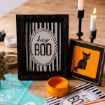 A framed “hey BOO” sign with black stripes and a framed black cat illustration on an orange background sit on a table with candles, orange paint, and Halloween-themed paper.