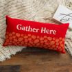 A red rectangular pillow with "Gather Here" text and pumpkin designs rests on a cream knit blanket next to an open book and a pair of eyeglasses on a wooden floor.
