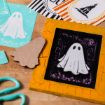 A handmade Halloween card featuring a white ghost on a black and orange background sits on a table with crafting supplies, including scissors, a cardboard ghost cutout, and other Halloween-themed cards nearby.