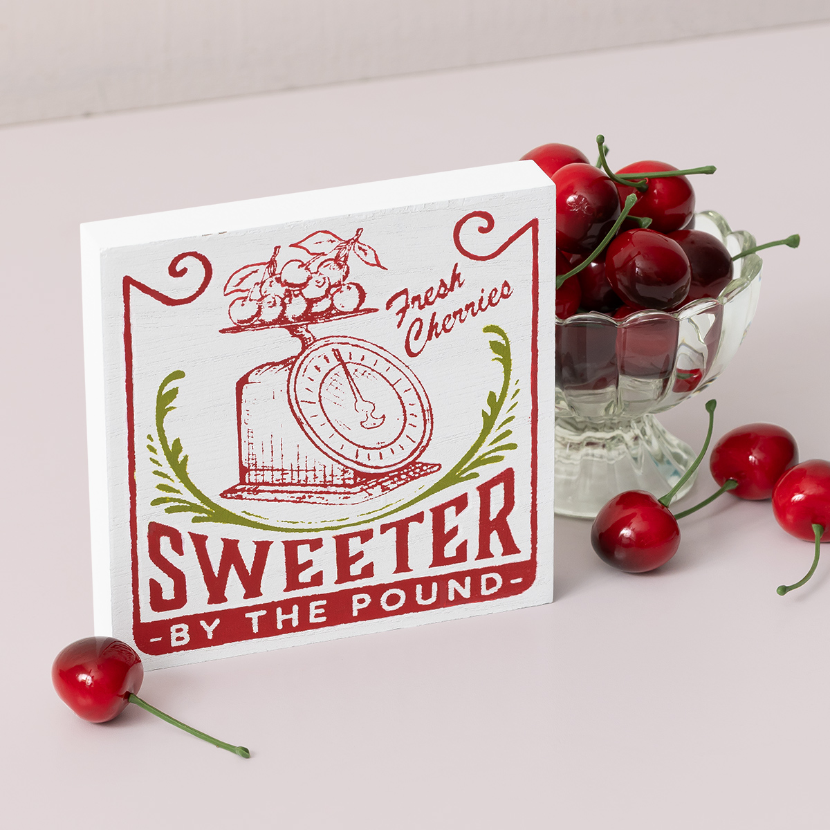 A square sign with an illustration of cherries on a vintage scale and the words “Fresh Cherries, Sweeter by the Pound.” A glass bowl filled with fresh cherries sits beside the sign on a light surface.