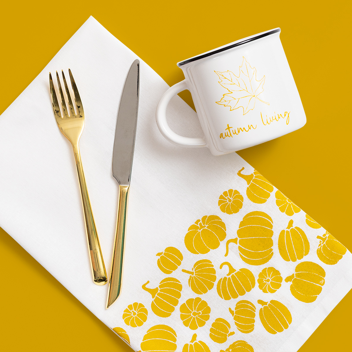 A white napkin with yellow pumpkin designs, a gold fork and knife, and a white mug with a maple leaf and “autumn living” text are arranged on a yellow background.