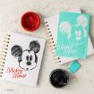 Two spiral notebooks with Mickey Mouse sketches on the covers, one white and one turquoise, are on a table with red and black paint jars and a turquoise eraser nearby. The Disney logo is visible.