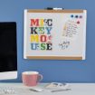 A whiteboard with "MICKEY MOUSE" in colorful letters, a weekly schedule, and magnets. A pink mug, keyboard, mouse, and a Mickey Mouse notepad rest on the desk below. Blue wall and part of a monitor are visible.