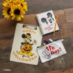 A clipboard with Mickey Mouse in a pumpkin costume, a Mickey Mouse notebook with a pencil, a pouch reading "Oh Boy!" with art supplies, and a vase of sunflowers on a wooden surface.