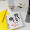 A notebook with a sketch of Mickey Mouse, a white water bottle with Minnie Mouse, a coaster with Donald Duck, a yellow folder, a pencil, and part of a laptop on a white desk.