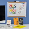 A whiteboard with colorful "MICKEY MOUSE" letters, a weekly planner, and magnets above a desk. Below are notebooks and cards with Disney characters. A computer is partly visible on the left.