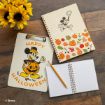 A clipboard and two notebooks with Mickey Mouse Halloween designs sit on a wooden table. A pencil rests on an open notebook, and a vase of yellow sunflowers is in the corner. © Disney.