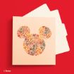 A card with a Mickey Mouse silhouette made of colorful autumn leaves, placed on a red background with two white envelopes partially visible behind it.
