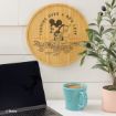 A round wooden plaque with an illustration of Mickey Mouse raking leaves and the text "Turning Over a New Leaf" hangs on a white wall above a laptop, a blue mug of coffee, and a potted plant.