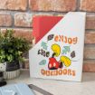 A white file holder with “Enjoy the Outdoors” text and a cartoon character's legs and shoes, sitting on a desk beside a plant, red file folders, and office supplies. Brick wall background.