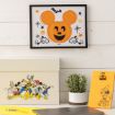 A framed Mickey Mouse pumpkin art hangs above a desk with a box featuring Disney characters, a small plant, and an orange notepad with a Minnie Mouse drawing.