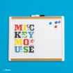 A whiteboard with colorful letters spelling "Mickey Mouse," each letter decorated with images of Mickey Mouse. Three magnets and a marker are on the board, and the background is bright blue.