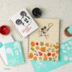 Spiral notebooks and cards featuring Mickey Mouse illustrations and autumn-themed designs, with jars of paint and art supplies on a light surface.