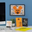A desk with Disney Halloween-themed art, including a framed Mickey pumpkin, Donald Duck, Minnie Mouse, and Goofy as a skeleton. Computer, plant, and yellow notebook also on the desk.