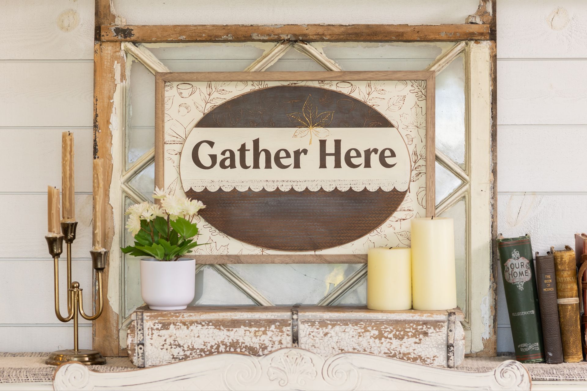 A decorative mantel displays a sign reading "Gather Here," flanked by candles, a white potted plant, books, and candlesticks, all arranged against a rustic wood and glass background.