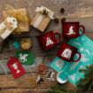 Three red mugs with Harry Potter designs, holiday gift tags, wrapped presents, pine branches, a stencil, and crafting materials are arranged on a wooden surface.