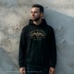 A man in a black "Order of the Phoenix Secret Society" hoodie stands against a textured, light-colored wall, looking slightly to the side.