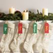 Four cream-colored knitted stockings hang from a white mantel, each labeled with a red or green tag reading Ron, Harry, Hermione, and Dobby. Candles and pine branches decorate the mantel above the stockings.