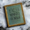 A framed green poster with ornate black text reads “Ministry of Magic” against a textured white and gray wall. There are small symbols and handwritten notes above and below the main text.