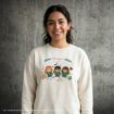 Smiling woman standing against a gray textured wall, wearing a cream sweatshirt with cute cartoon depictions of three young characters in festive holiday outfits and decorative elements.