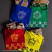 Four colorful tote bags—blue, green, red, and yellow—each featuring a Hogwarts house crest. Each bag holds wrapped gifts decorated with gold ribbons. The bags are arranged in a square on a dark surface.