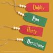 Four wooden tags in red and green, each with a name: Dobby, Ron, Harry, and Hermione, written in yellow or white, with string and wooden beads attached, arranged diagonally on a mustard yellow background.
