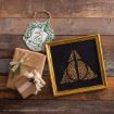 Two brown paper gift boxes, a green Slytherin ornament, and a gold-framed art piece featuring words forming the Deathly Hallows symbol are arranged on a wooden surface.