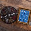 A round Platform 9¾ sign with magical illustrations, next to a framed blue Harry Potter 25th anniversary print with white silhouettes of characters and magical symbols on a wooden surface.