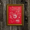 A framed red poster on wood shows Christmas-themed Harry Potter drawings with white text reading “All I Want for Christmas” and various related doodles like a scarf, wand, owl, sweater, and socks.