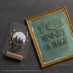 A framed “Ministry of Magic” print sits next to a Harry Potter-themed pin featuring a silhouette of Hogwarts with gold stars and symbols on a gray backing card.