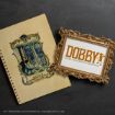A vintage Harry Potter notebook lies beside a gold ornate frame with “DOBBY has come to save Harry Potter and his friends” written inside on a dark background.