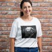 A smiling woman stands in front of a brick wall, wearing a white shirt with a "Magic Spells & Potions" design featuring silhouettes of witches on brooms.
