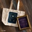 A beige tote bag with a mystical design and a framed “Midnight Margaritas” cocktail recipe are displayed on a wooden floor. Both items have a magical, witch-themed aesthetic.