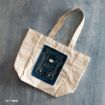 A beige tote bag lies on a textured surface, featuring a dark blue design with the words “Witch and Wizard” and illustrated magical elements on its front.
