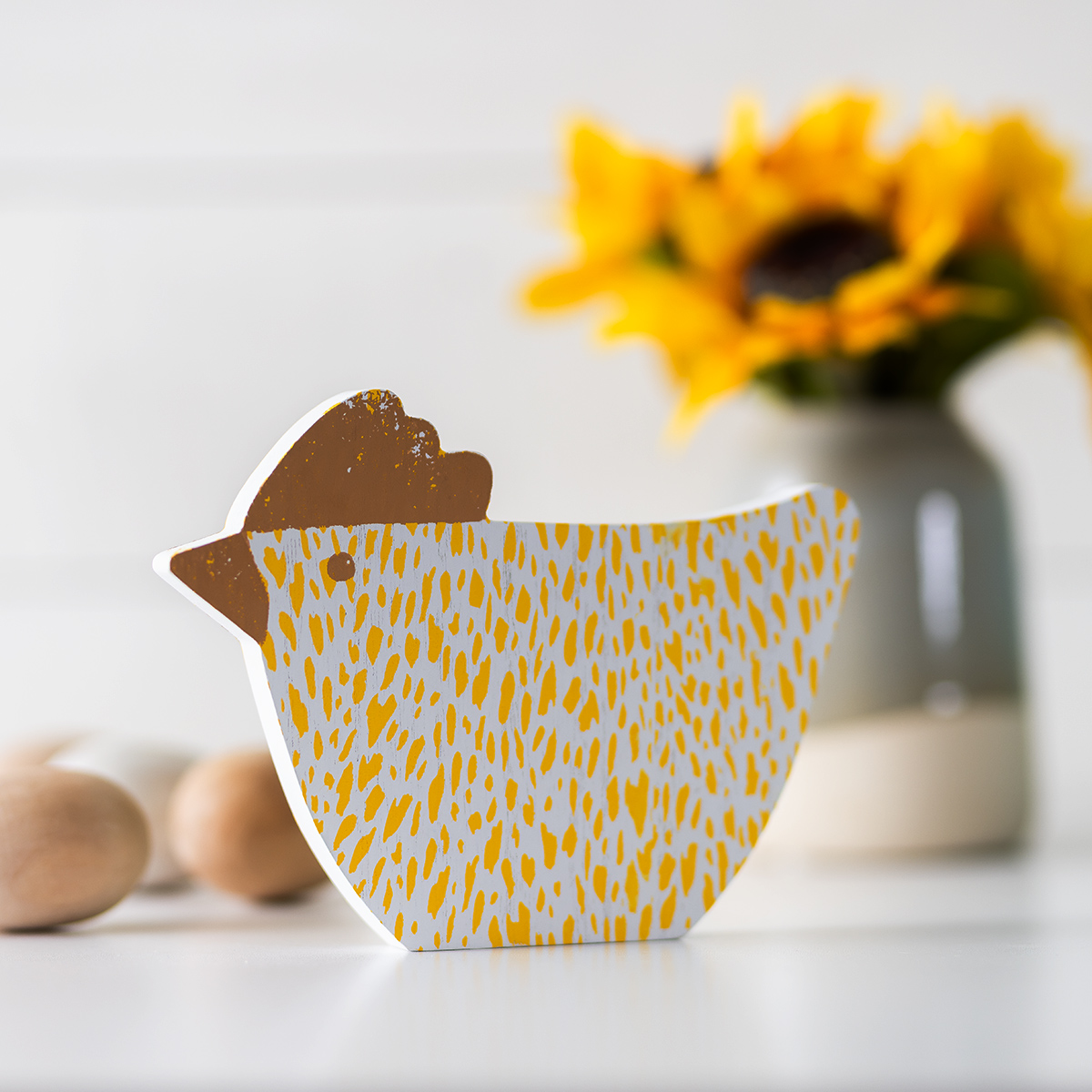 A wooden chicken figurine with yellow speckles stands on a white surface. A vase with sunflowers and blurred objects are in the background.