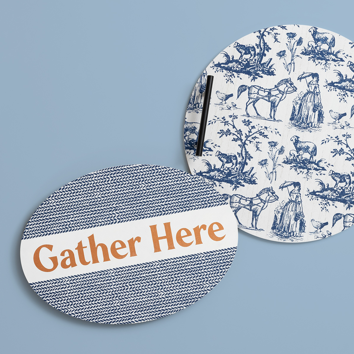 Two round placemats on a blue background: one with blue patterns and “Gather Here” in large orange letters, the other with blue illustrations of people, horses, and trees.