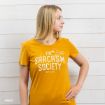 A woman in a mustard yellow t-shirt with “The Sarcasm Society” and a cartoon character stands against a white wooden wall, looking to the side with one hand on her hip.