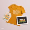 A mustard yellow “Sarcasm Society” T-shirt, a Peanuts-themed tag, and a framed illustration of Peanuts characters in a pumpkin patch are arranged on a light pink surface.