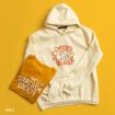A cream hooded sweatshirt with “Sweater Weather” and a Peanuts design next to a mustard yellow T-shirt that says “The Sarcasm Society” with Peanuts characters, on a yellow background.