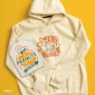 A cream hoodie with “Sweater Weather” and Peanuts characters sits next to a pumpkin-shaped Peanuts sign on a solid yellow background.