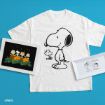 A white t-shirt with a large Snoopy print is placed on a blue surface, flanked by two framed Peanuts illustrations: one of kids in a pumpkin patch and another with Snoopy and Woodstock hugging.